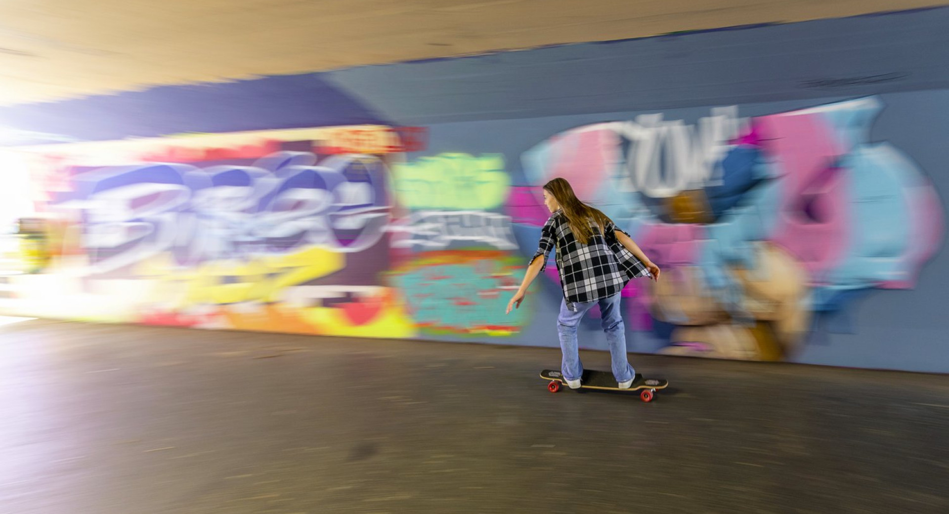 A young girl skateboards in front of colorful graffiti. A young girl skateboards in an underpass.