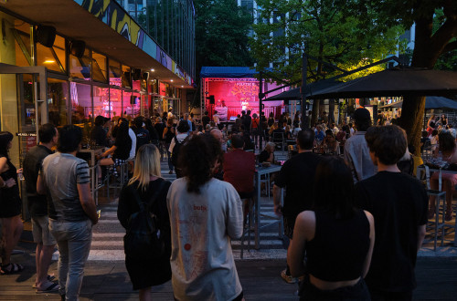 People sit at café tables, chatting and listening to a musical performance.