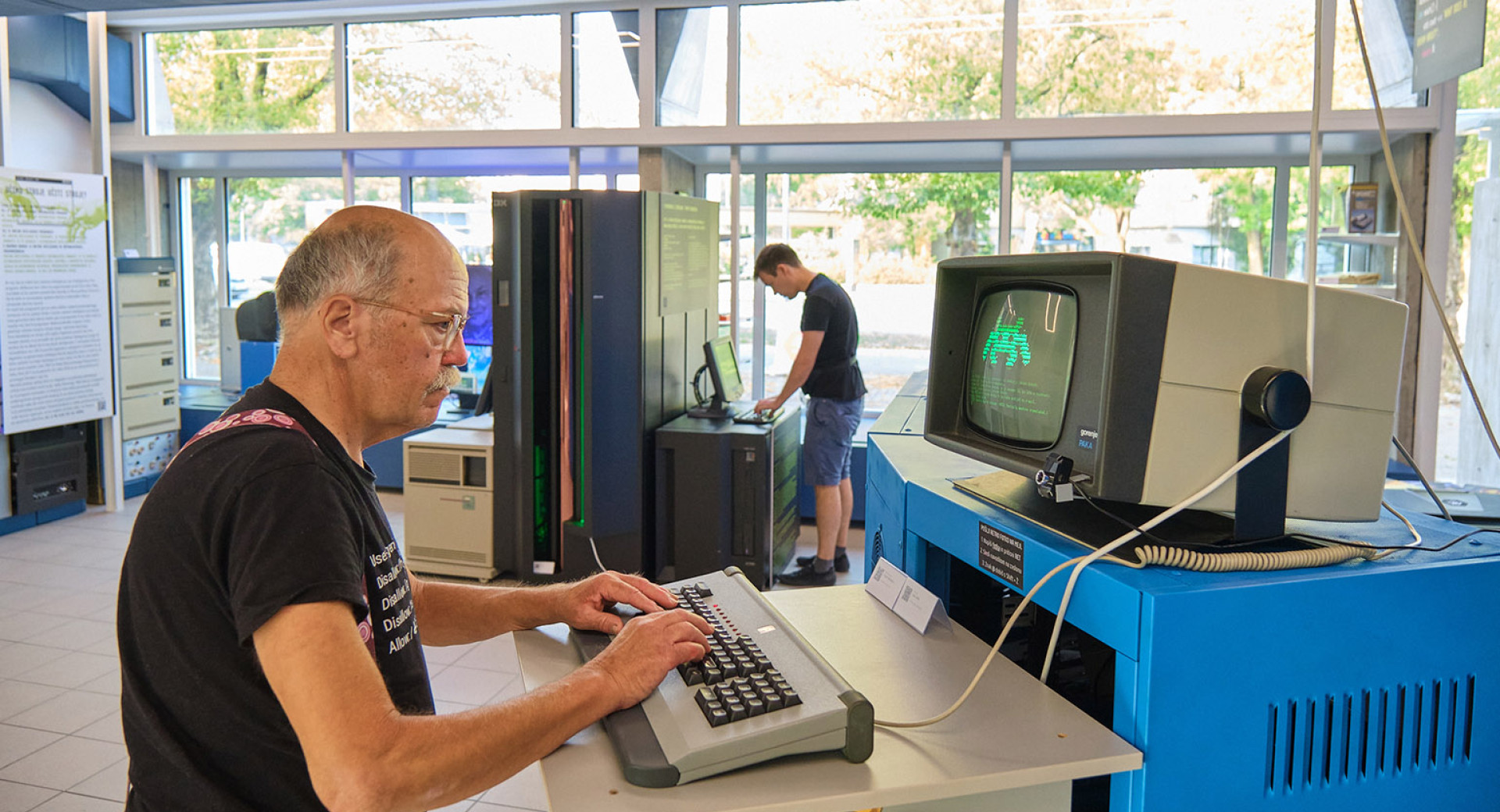 An older man types on a computer. A vintage computer is displaying a game