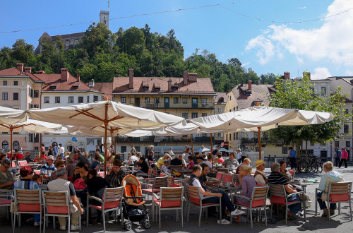 Obiskovalci na zunanji terasi pod senčniki. V ozadju Ljubljanski grad.