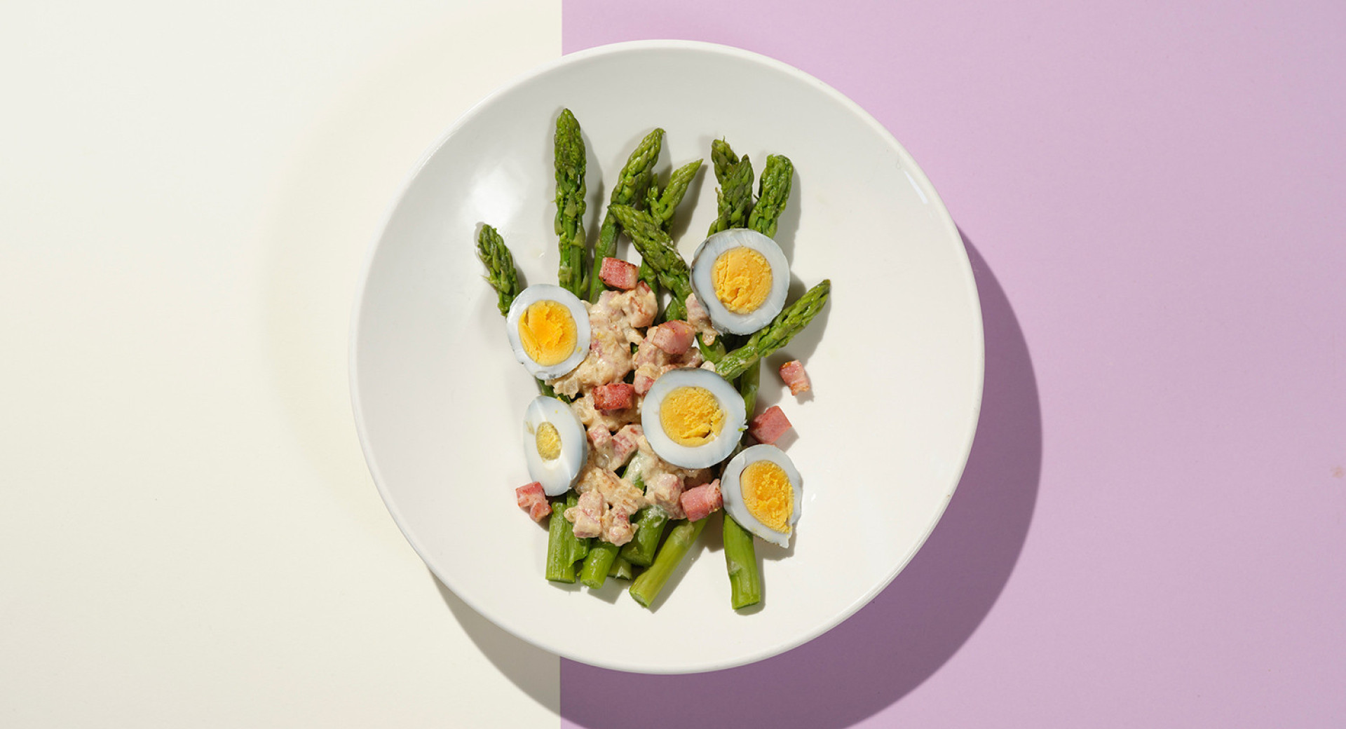 A white plate on a beige and violet background. The plate features neatly presented asparagus with chopped hard-boiled eggs, sprinkled with prosciutto.