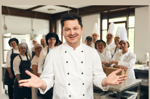 A man in a white chef's uniform, his team in the background with their arms spread wide.