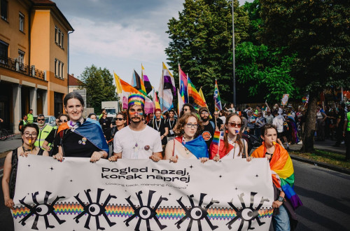 Moski in zenske drzijo v rokah napis Pogled nazaj, korak naprej. V ozadju mavricne zastave.