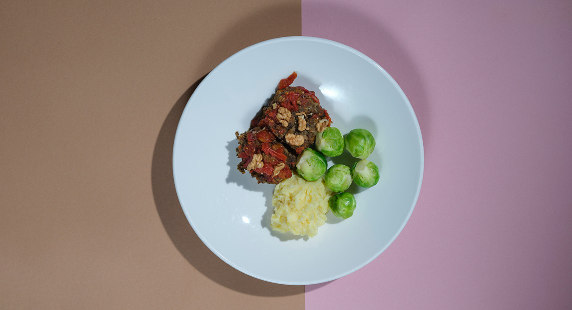 A white plate on a brown and pink base. On the plate, there is a neatly arranged roast with lentils and walnuts, alongside Brussels sprouts and mashed potatoes.