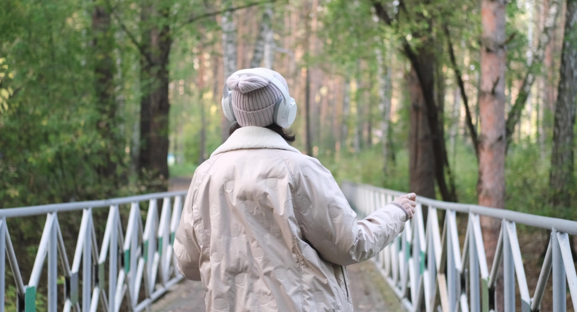 A woman walks across a white bridge in the middle of a forest. A woman is wearing headphones.