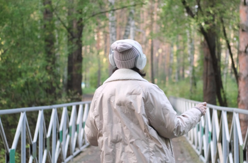 A woman walks across a white bridge in the middle of a forest. A woman is wearing headphones.