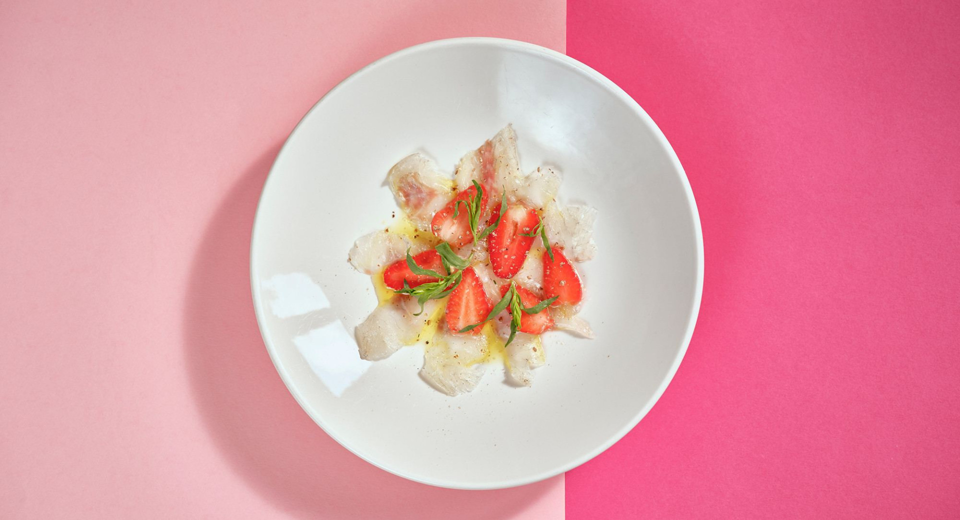 A white plate on a light and dark pink base. On the plate, neatly arranged fish, decorated with strawberries and greens.