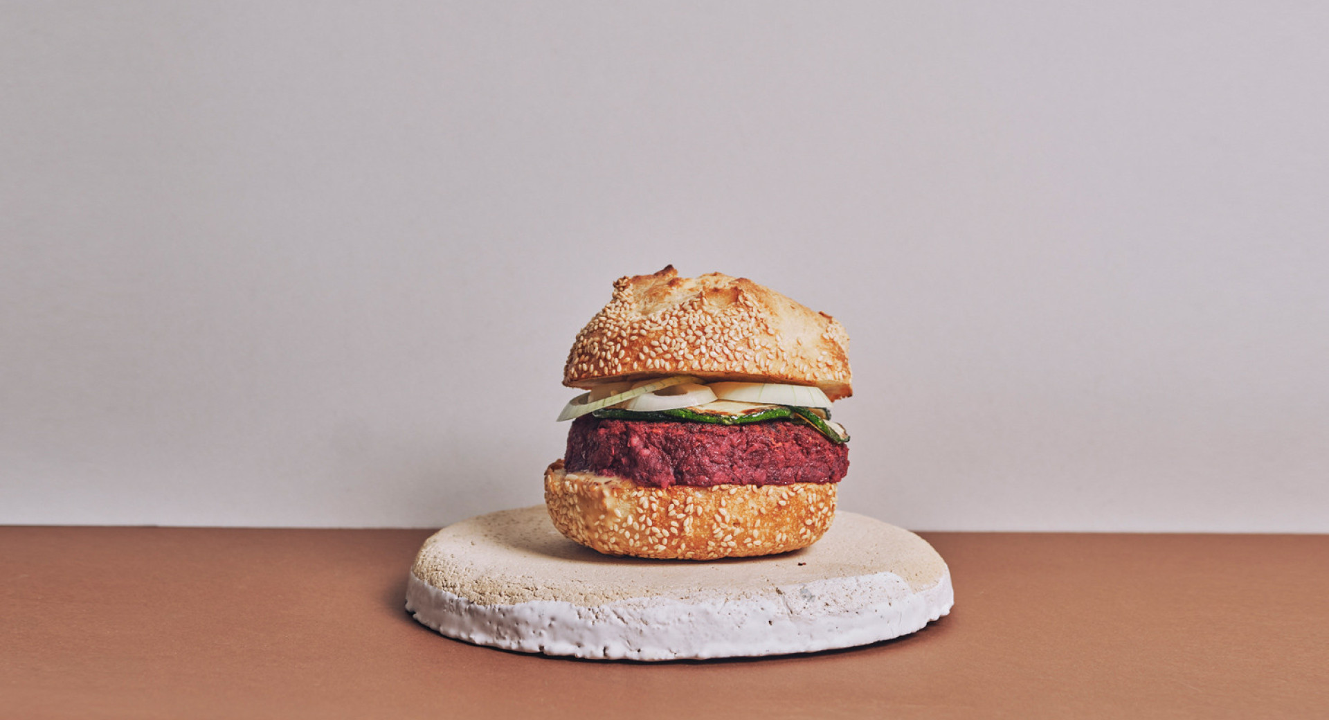 kamnit kroznik na rjavi mizi, belo ozadje. na krozniku predstavljen domac burger, z debelim zrezkom in zelenjavo med rezinama zemljice. 