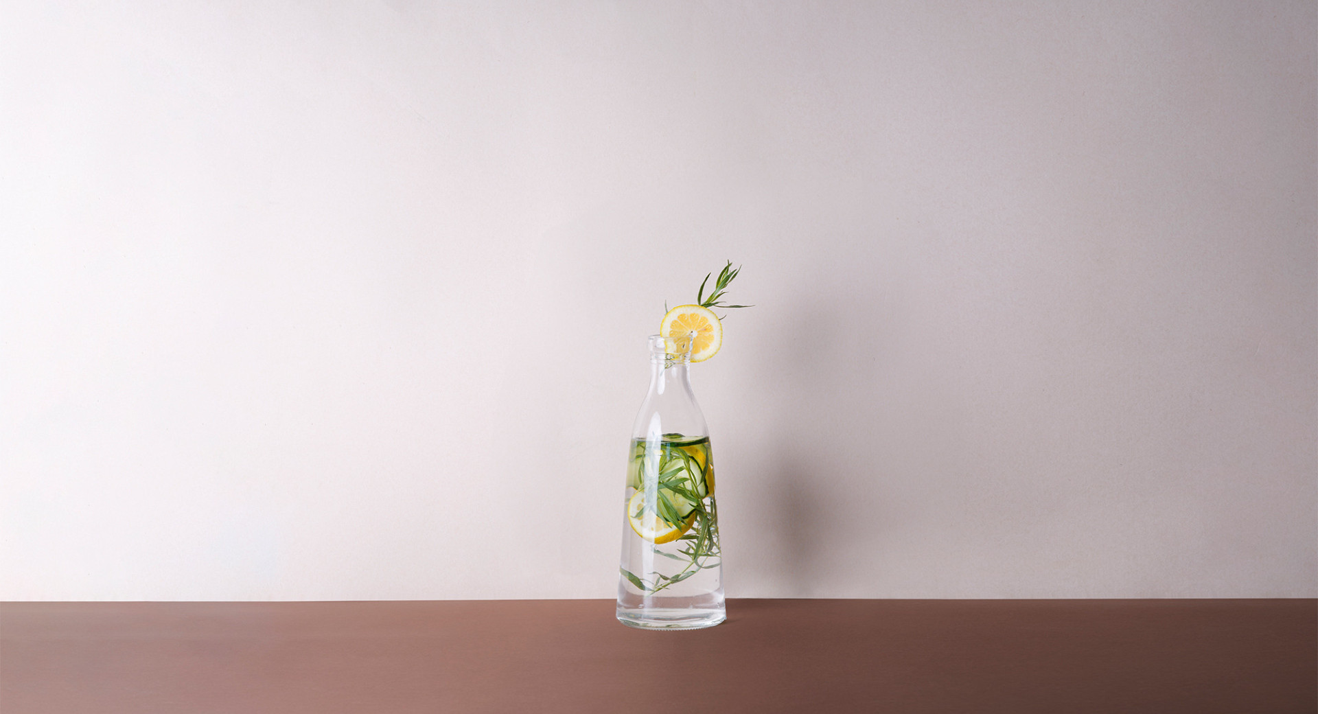  A glass jar on a brown table, with a white background. Inside the jar, there's lemonade with sprigs of mint and slices of lemon.