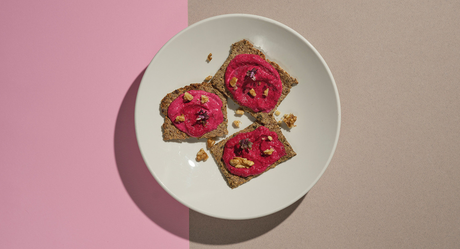 A white plate on a pink and brown background. Neatly arranged brown crackers with pink sauce are placed on the plate. Pumpkin seeds are sprinkled on top for decoration.