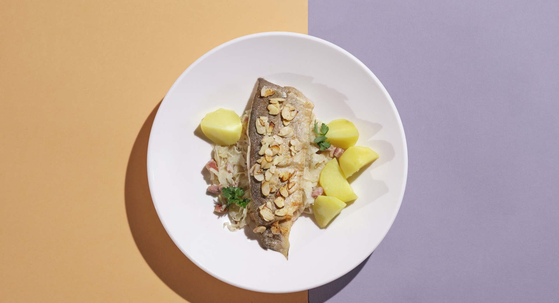 A white plate on an orange and purple base. On the plate, there's a fish fillet and potatoes in pieces, garnished with chopped parsley and almonds.