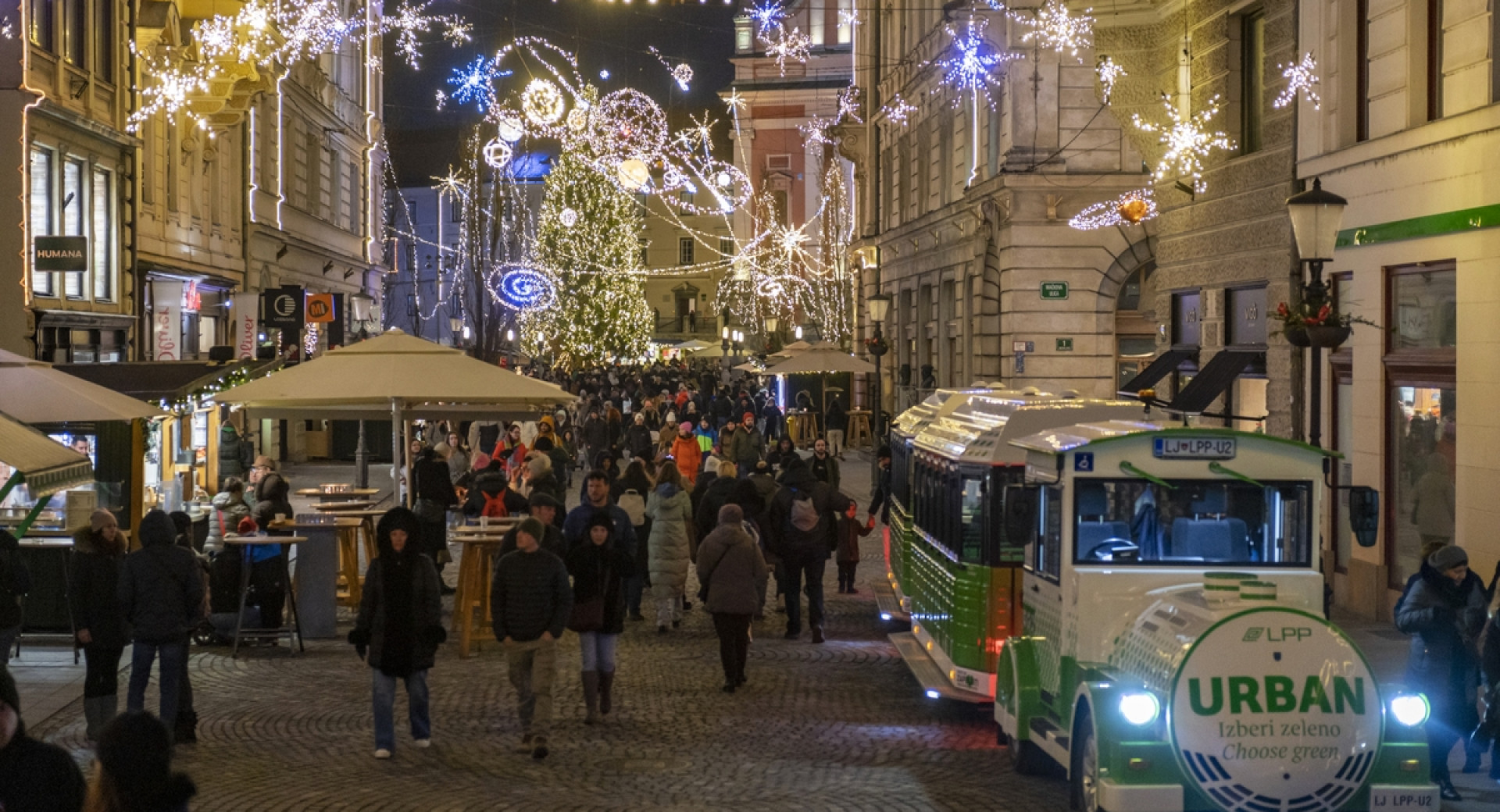 Ljudje v novoletno okrašenem mestu, ob strani belo-zelen vlakec.