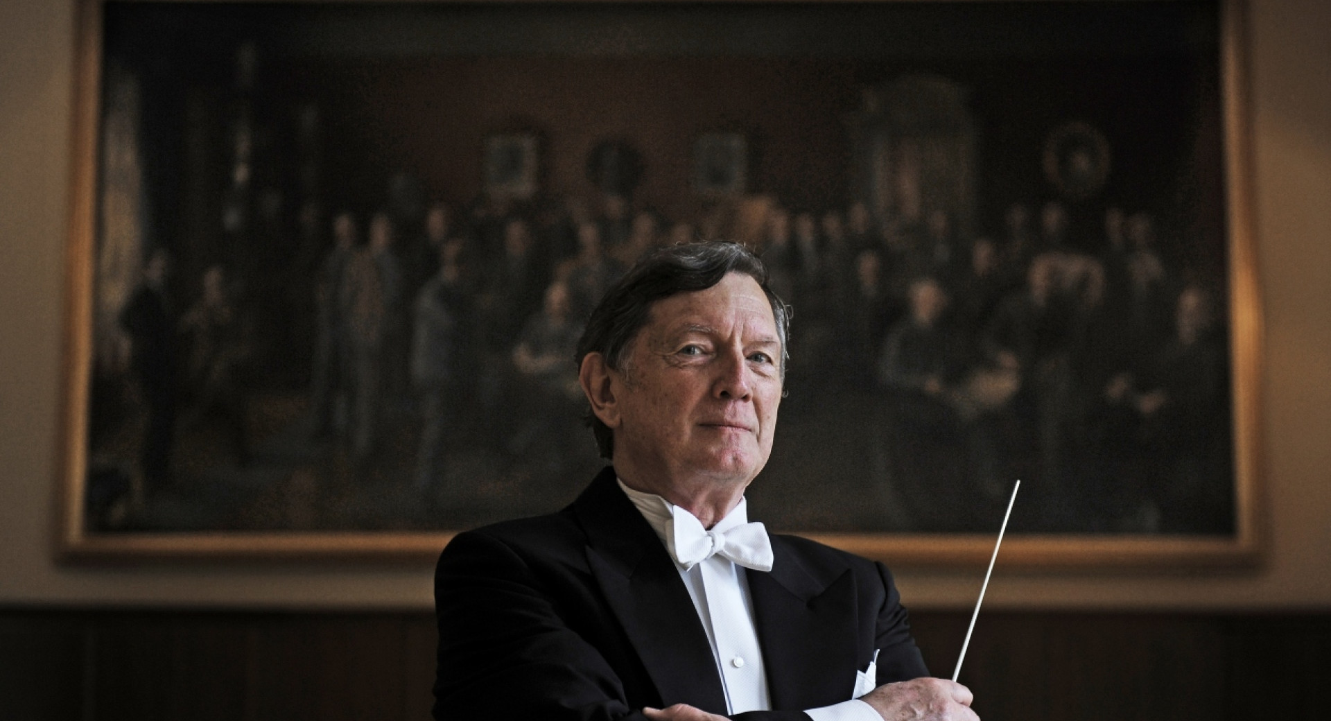 A man in formal attire with a bow tie and a conductor's baton confidently stands in front of an artwork in an elegant setting.