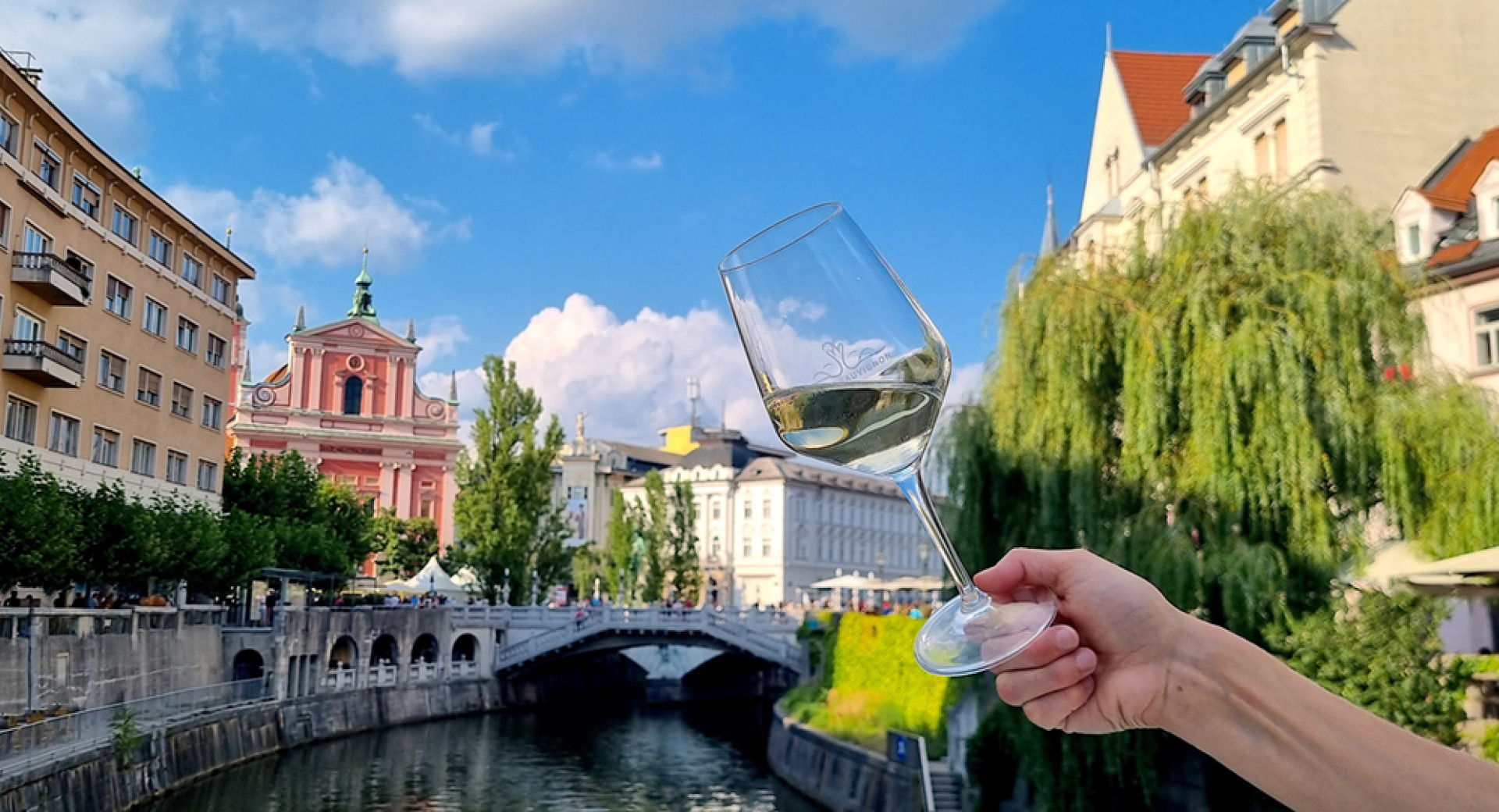 Oseba drži kozarec belega vina z razgledom na zgodovinsko mestno jedro Ljubljane