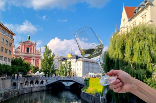 Oseba drži kozarec belega vina z razgledom na zgodovinsko mestno jedro Ljubljane