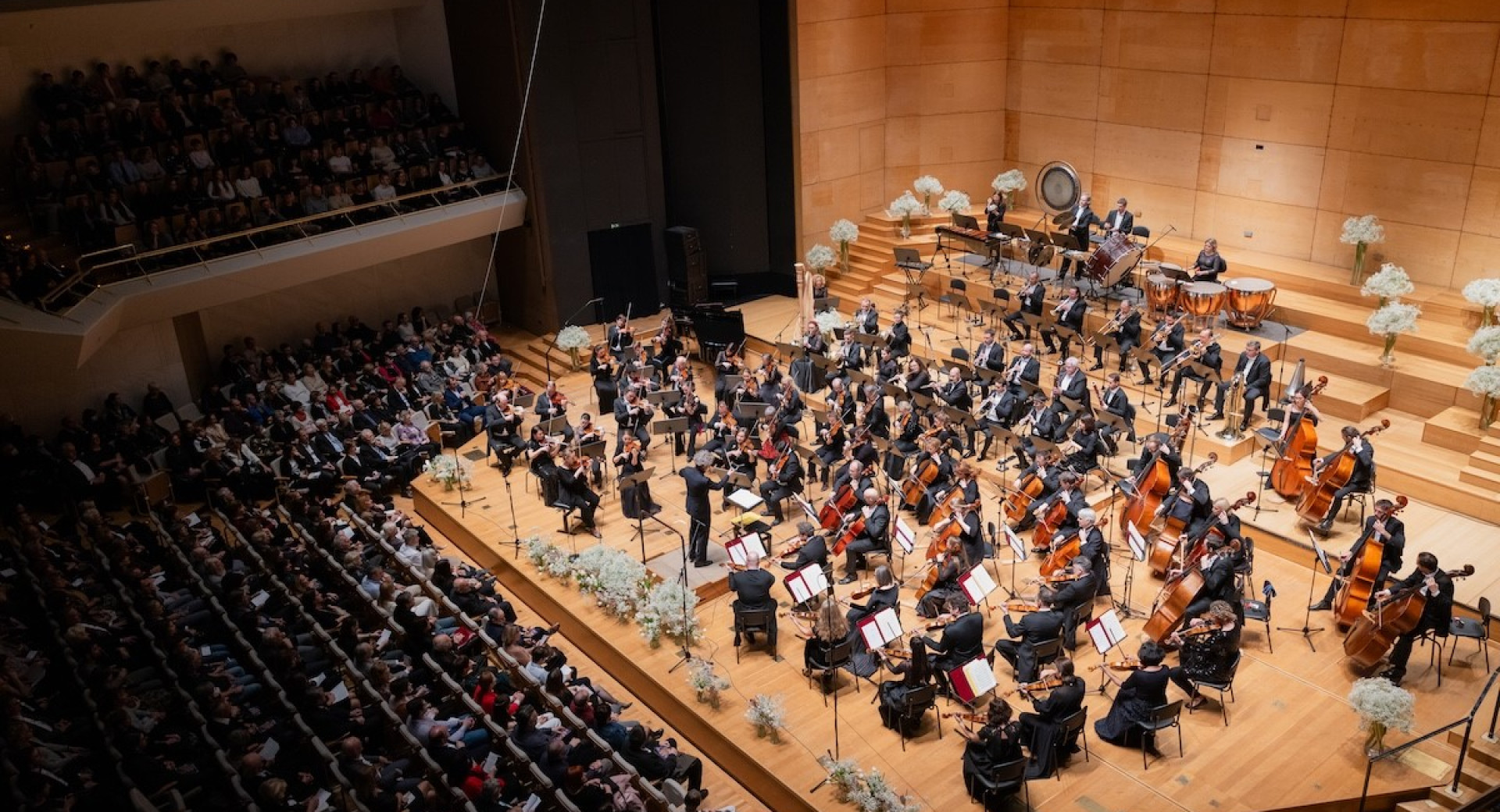 The philharmonic orchestra is performing on a wooden stage in front of a crowd in the hall.