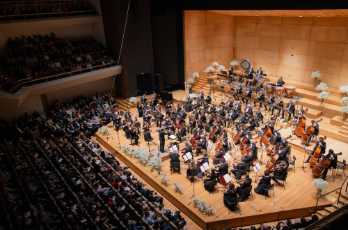 Filharmonični orkester na lesenem odru nastopa pred množico ljudi v dvorani.