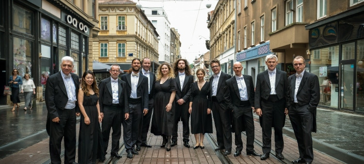 Skupina ljudi v elegantnih oblacilih stoji na tramvajskih tirih v mestnem srediscu, obdana z mesanico zgodovinskih in sodobnih stavb.