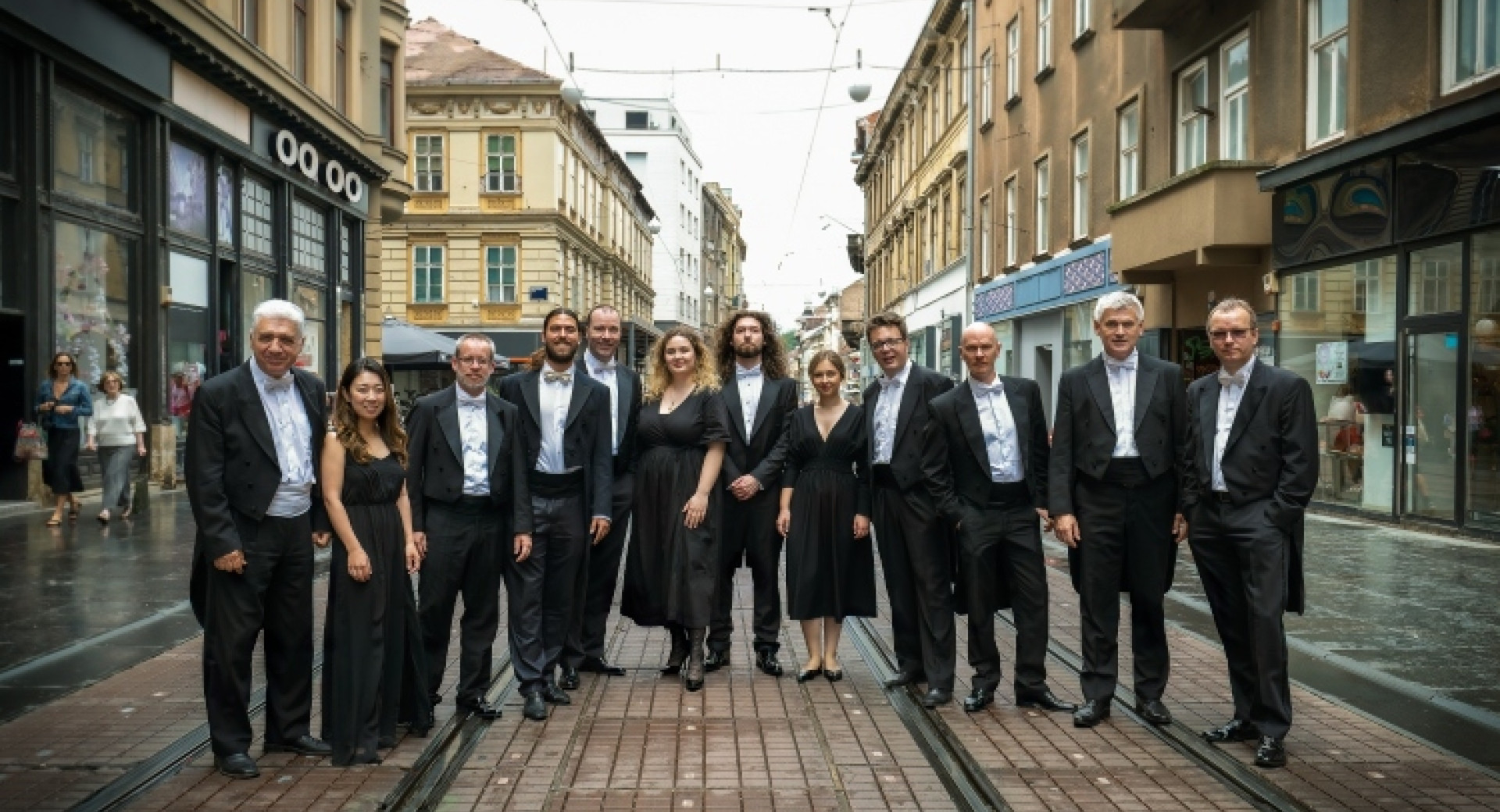 Skupina ljudi v elegantnih oblacilih stoji na tramvajskih tirih v mestnem srediscu, obdana z mesanico zgodovinskih in sodobnih stavb.