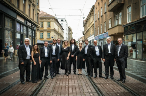 Skupina ljudi v elegantnih oblacilih stoji na tramvajskih tirih v mestnem srediscu, obdana z mesanico zgodovinskih in sodobnih stavb.