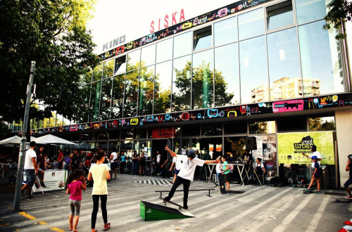 People in front of a large glass building. A boy is skateboarding in front of the building.