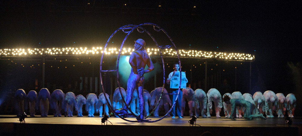 People in white clothing are arranged on the stage. An acrobat in a white costume is in the middle of a large iron ring in the center of the stage.