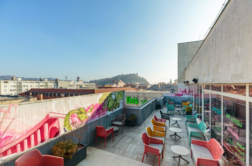 A terrace with colourful chairs and small tables offers a view of the cityscape. The concrete wall is covered in vibrant graffiti, and in the background, under a clear blue sky, you can see a hill with a castle.