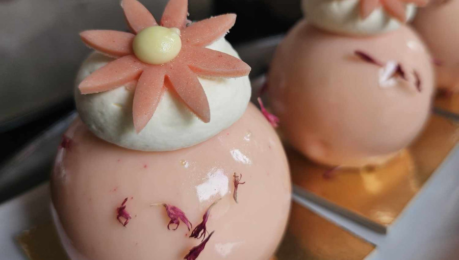 Desserts consisting of a shiny pink ball with a floral decoration, set on a gold plate. In the background there are white chairs and blurred furniture.