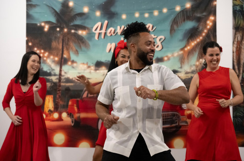 A group of people dance in front of a tropical backdrop of palm trees and fairy lights. The mood is cheerful, with three women in red dresses dancing in the background, and a man in a white shirt in the foreground, who is also smiling and dancing.