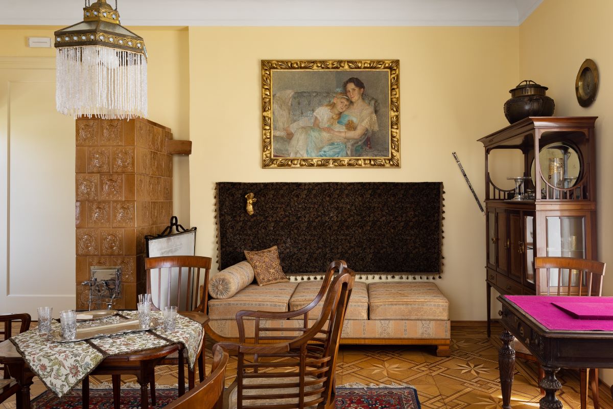 An elegant interior featuring wooden furniture, a ceramic stove on the left, and an art piece in a golden frame hanging above a cushioned bench, all in warm earthy tones.