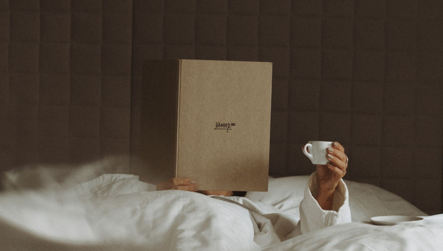 A person, bundled in a white blanket is laying in bed, holding a small white cup and covering their face with a large book.