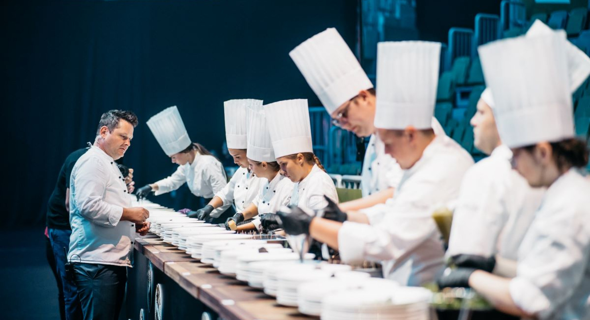 Young chefs prepare dishes while being observed by a master chef.