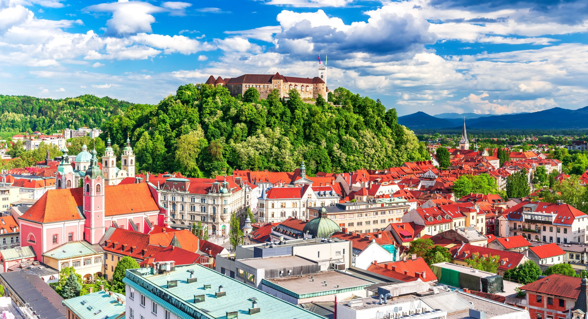 A view of rooftops of a city. Green hill with a castle on top reigning over the town. Blue sky.   