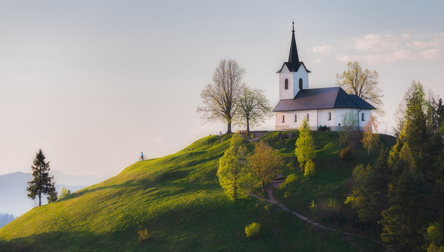 A church on top of a small hill. A cyclist on the hillside.