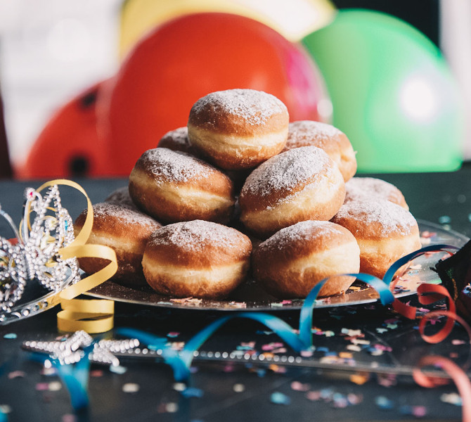 A large pile of donuts on a plate. The plate is on a black table decorated with confetti, colorful ribbons, and a crown. In the background, colorful balloons are out of focus.