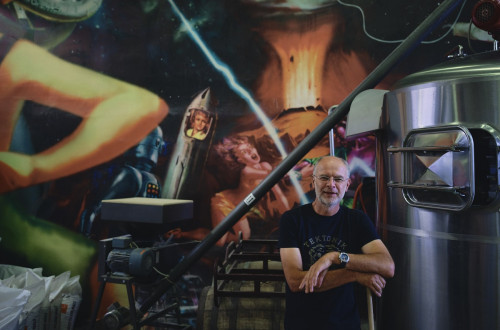 A man in a black t-shirt is standing in front of a wall with graffiti, and there is a large metal barrel next to him.