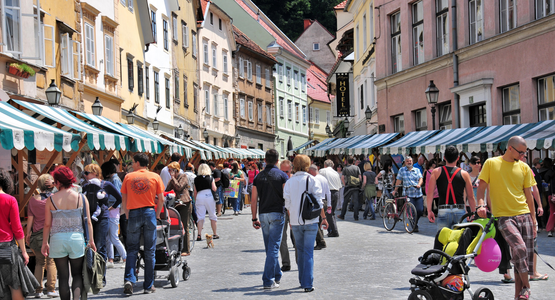 Zivahna ulica z barvitimi stavbami in stojnicami. Ljudje se sprehajajo in si ogledujejo stojnice, nekateri potiskajo otroske vozicke.