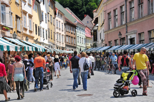 Zivahna ulica z barvitimi stavbami in stojnicami. Ljudje se sprehajajo in si ogledujejo stojnice, nekateri potiskajo otroske vozicke.