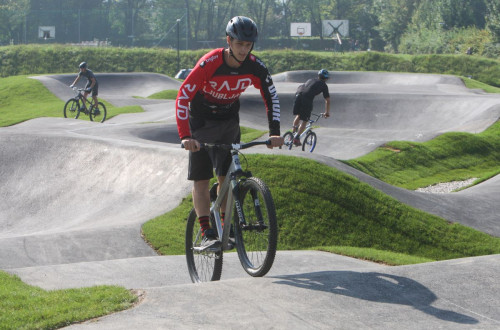 Mladenic na kolesu na pumptrack stezi, obdan z zelenimi gricki, v ozadju pa so vidni se drugi kolesarji, ko premagujejo valovite poti.