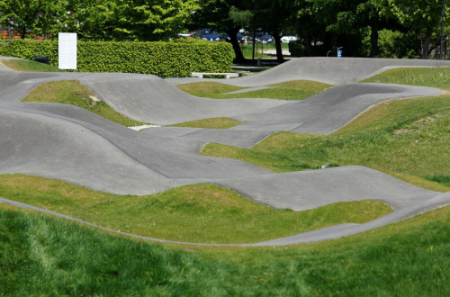 Pumptrack steza, obdana z zelenico in drevesi, ponuja valovite asfaltne poti za rekreativne kolesarske podvige.