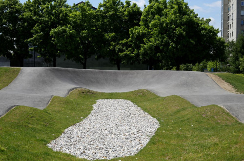 Pumptrack steza z znacilnimi valovitimi potezami, obkrozena z zeleno travo in posuta z belimi kamencki, obdana z drevesi v ozadju.