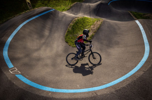 Pumptrack steza s poudarjenimi modrimi linijami, na kateri kolesar v zascitni opremi premaguje zavoj.