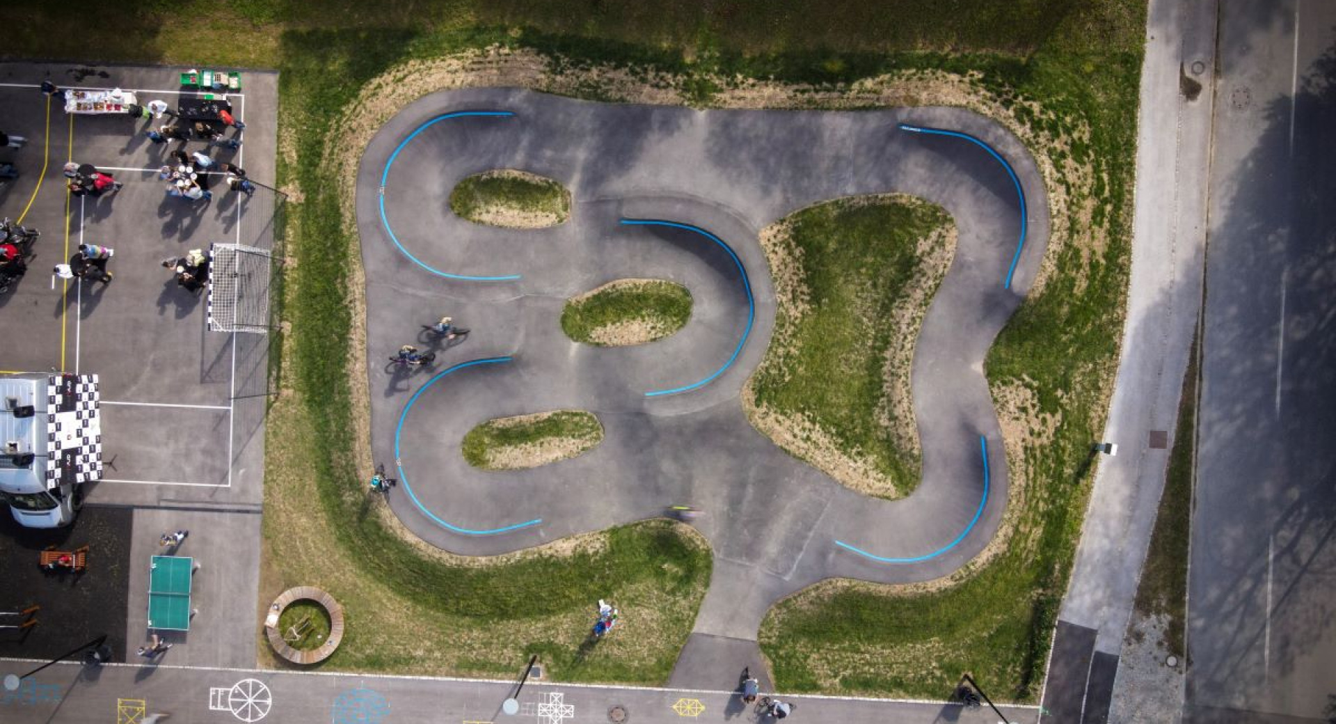 Zracni posnetek pumptrack steze s tekocimi zavoji in modrimi oznacevalnimi linijami, obkrozene z zeleno travo in spremljevalnimi povrsinami za gledalce.