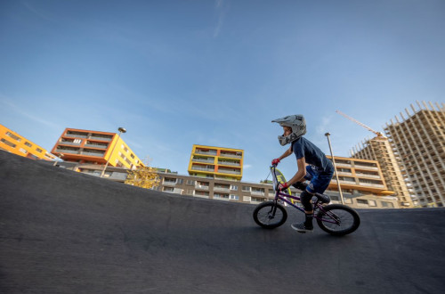 Fotografija prikazuje mladega kolesarja z BMX-om na pumptrack stezi, obdanega z modernimi, pisanimi stanovanjskimi bloki in gradbenim zerjavom v ozadju.