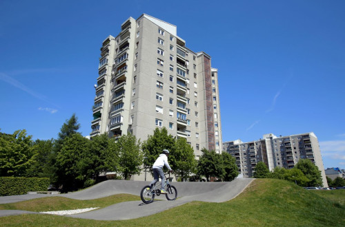 The photo shows a cyclist on a pump track surrounded by greenery, with tall residential buildings in the background under a clear blue sky.