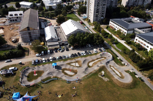 Pumptrack steza iz zraka z okoliskimi stavbami, avtomobili in dogodkom, kjer se zbirajo obiskovalci pod sotori.