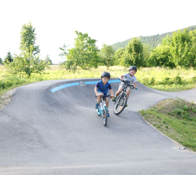 Otroci med voznjo na pumptrack stezi, obdani z naravo v poletnem vzdusju.