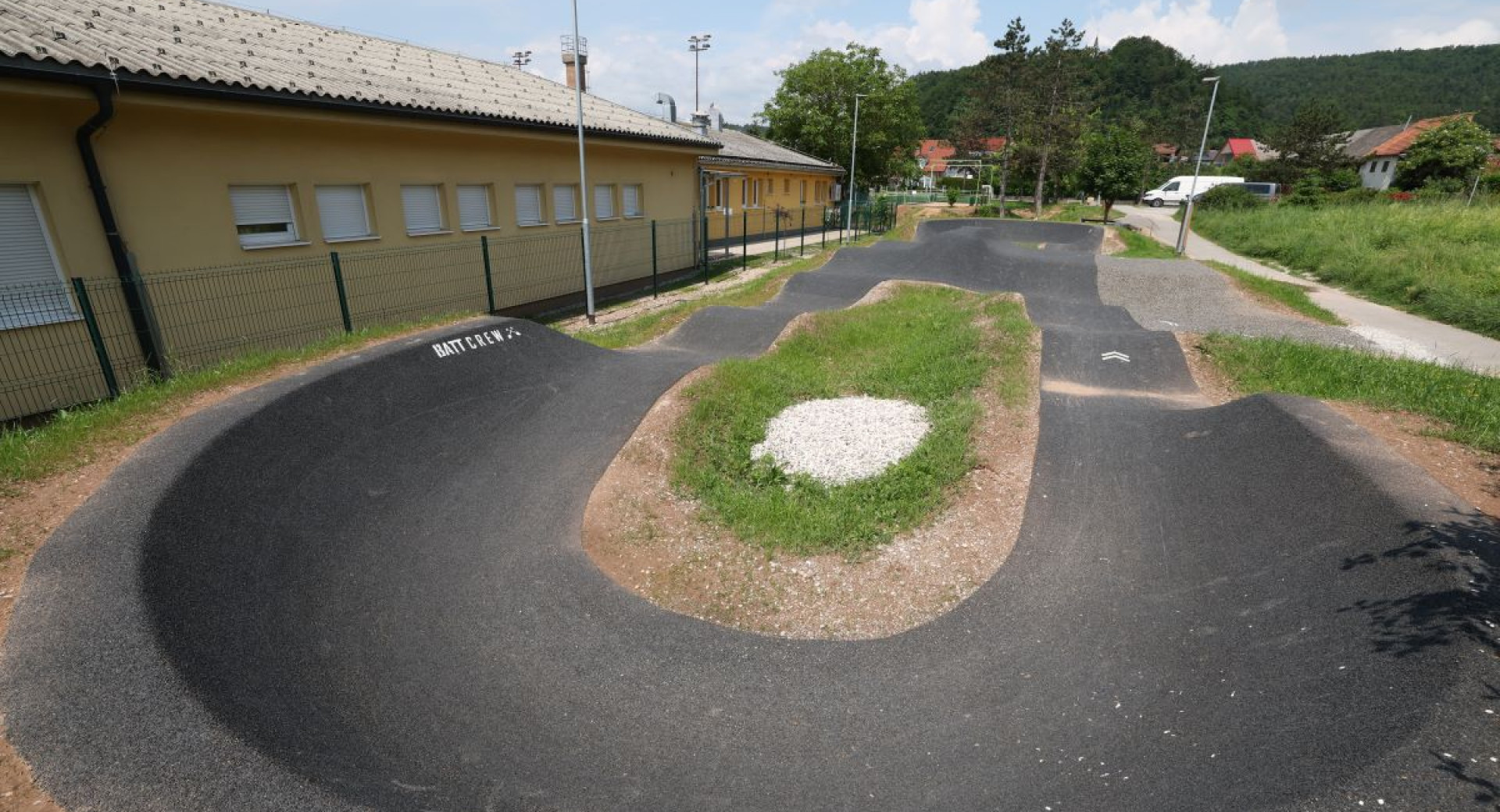 Pumptrack steza ob stavbi v soncnem vremenu, obkrozena z zelenjem in razgibano povrsino za kolesarjenje.