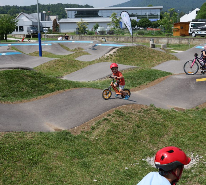 Otrok na poganjalcku se vozi po valovitem terenu pumptrack poligona, obdan z naravo in kolesarsko opremo.