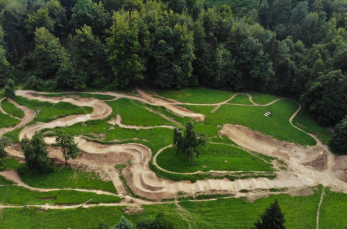 Pumptrack poligon, umescen v naravno okolje, je sestavljen iz valovite proge z ovinki, ki se razteza ob robu gozda in ponuja možnost za rekreativno kolesarjenje.